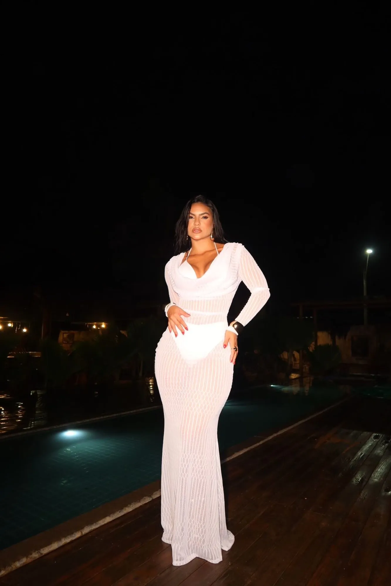 Modelo em deck iluminado à beira da piscina usando vestido longo branco de tricô vazado com mangas longas